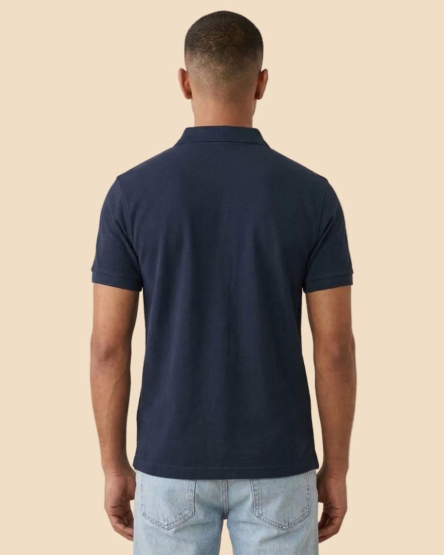 A man viewed from behind wearing a navy blue short-sleeve polo shirt and light wash jeans, standing against a plain beige studio background.
