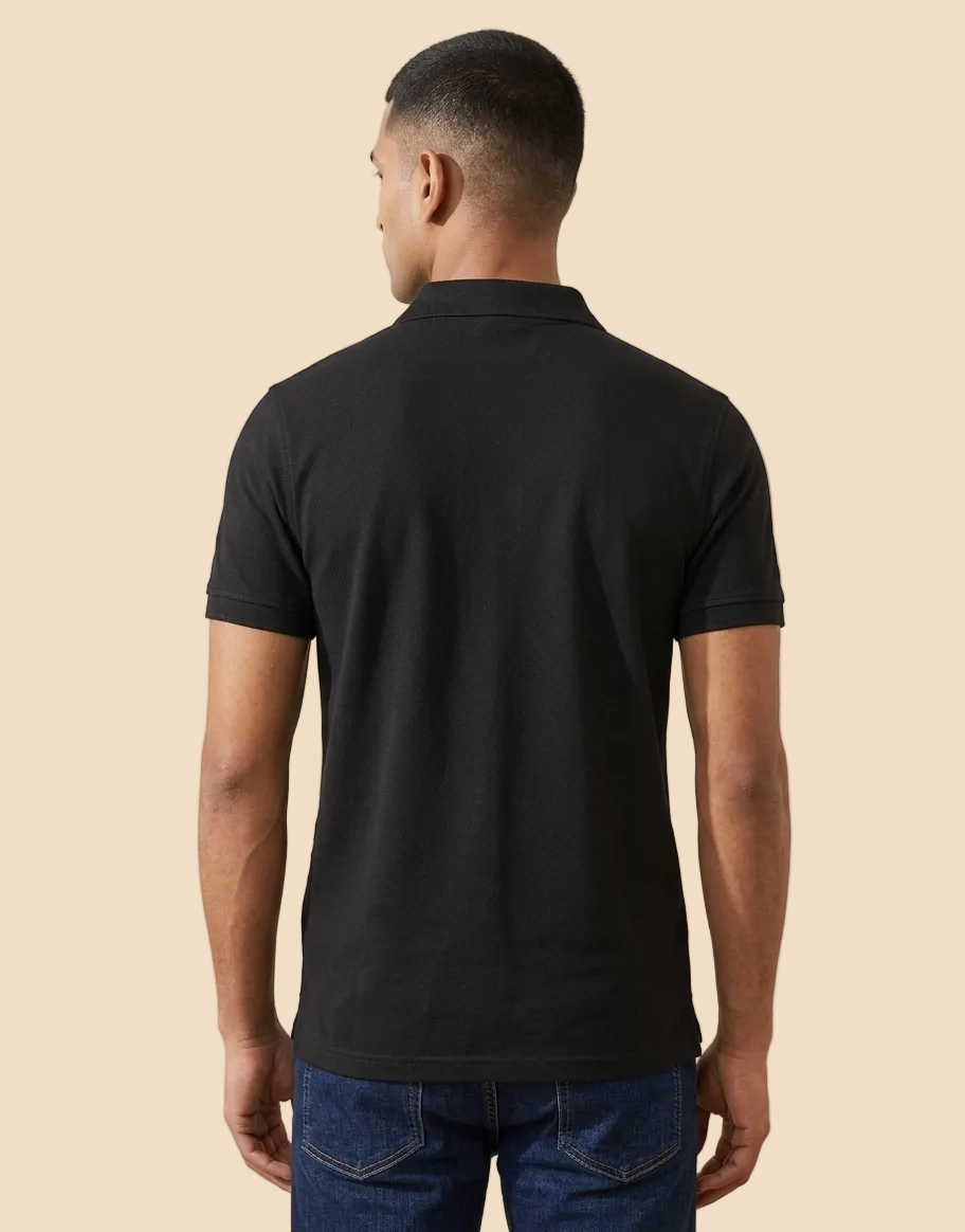 A man viewed from the back wearing a plain black short-sleeve polo shirt and blue jeans, standing indoors against a light beige wall.