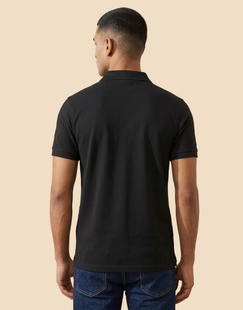 A man viewed from the back wearing a plain black short-sleeve polo shirt and blue jeans, standing indoors against a light beige wall.