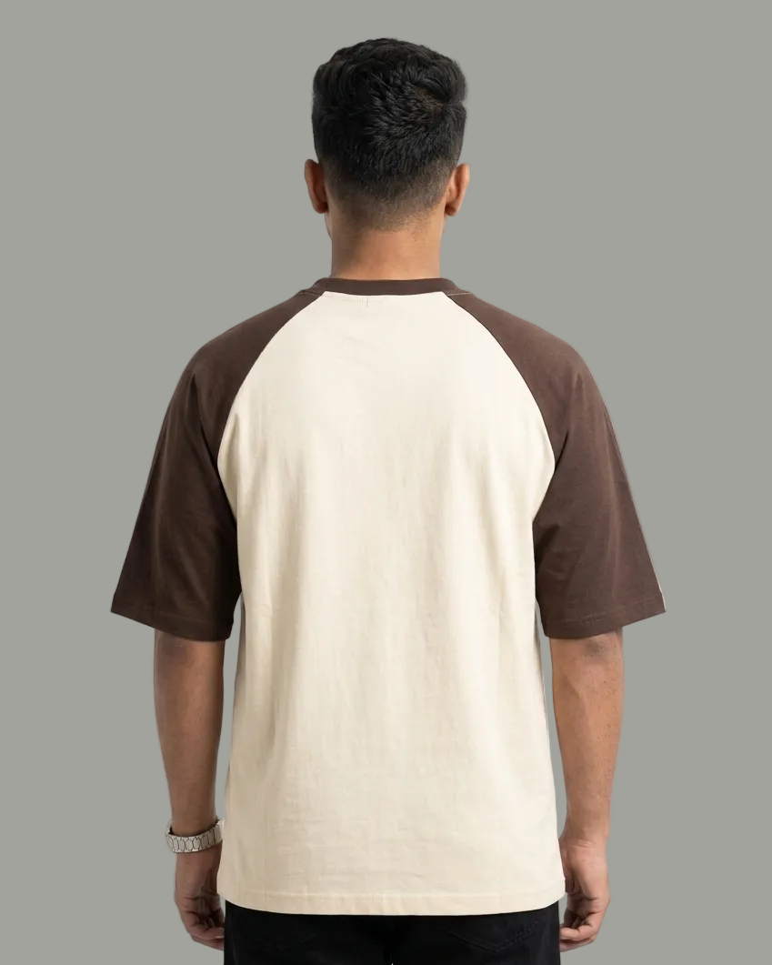 A man viewed from behind wearing a short-sleeve raglan t-shirt with a cream-colored body and dark brown sleeves and collar, set against a plain white studio background.