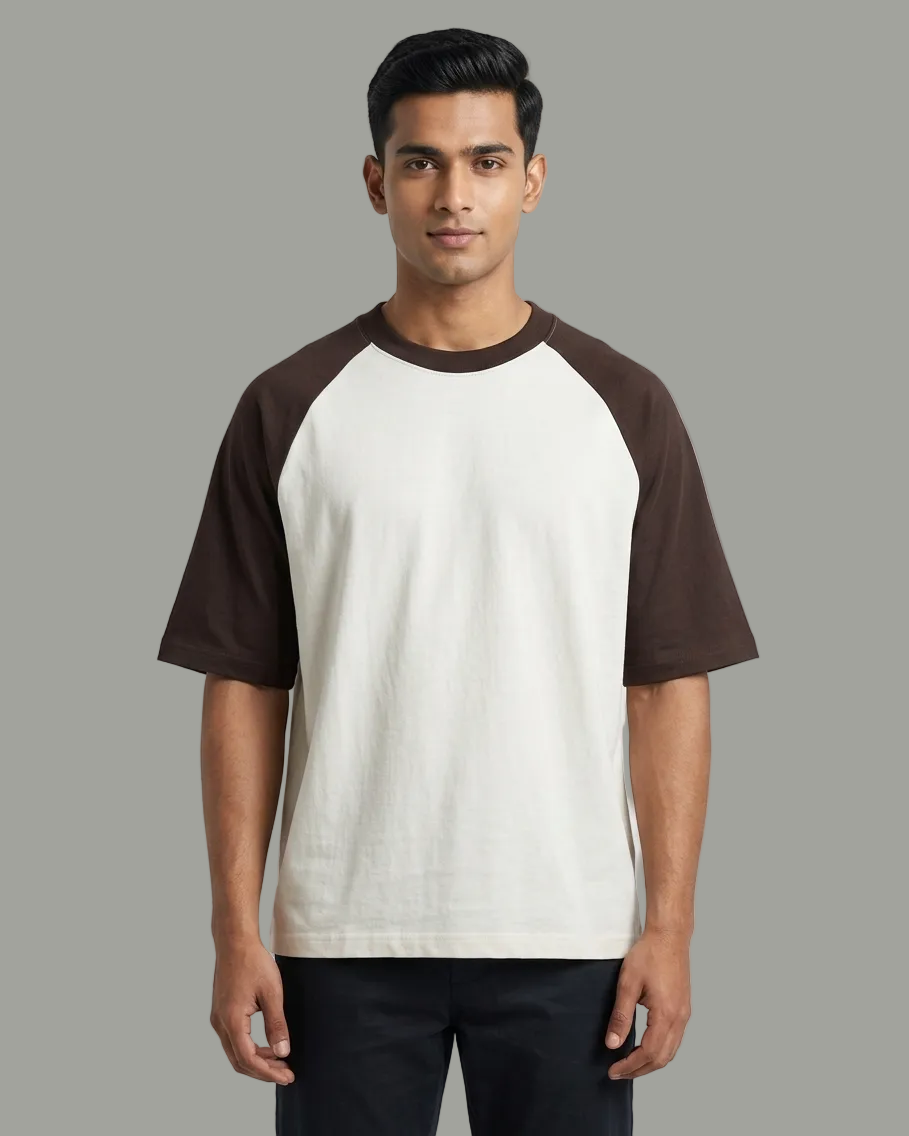 A man wearing a short-sleeve raglan t-shirt with an off-white body and dark brown sleeves, paired with dark pants, against a light gray studio background.