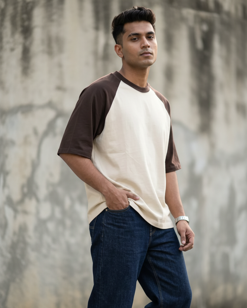 A man wearing a cream and brown raglan t-shirt with dark blue jeans, standing against a concrete wall.