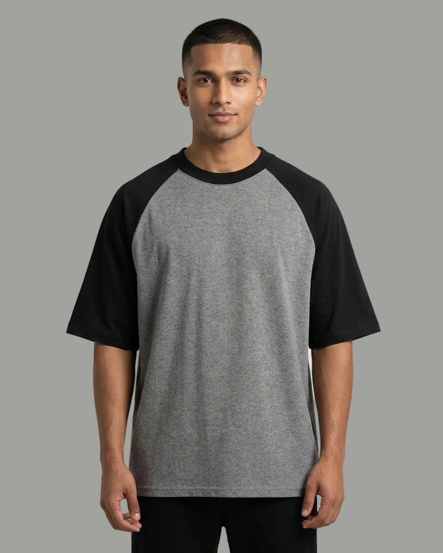 A man wearing a short-sleeve raglan t-shirt with a heather gray body and black sleeves, standing against a light gray studio background.