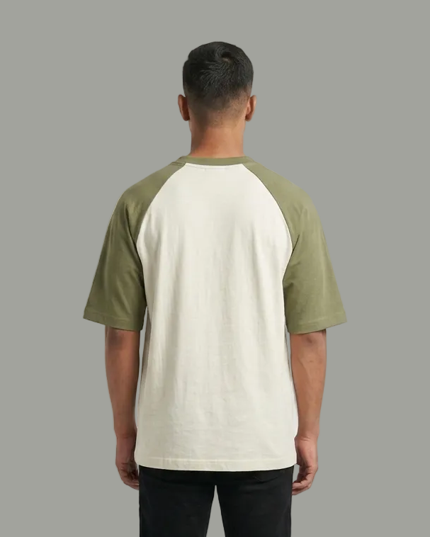 A man viewed from the back wearing a raglan-style t-shirt with an off-white body and olive green sleeves, paired with black pants and white sneakers, against a light gray studio background.