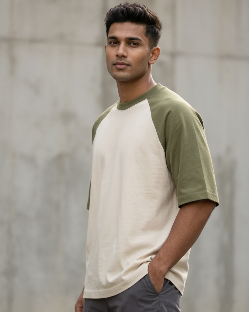 A man wearing a cream and olive green raglan t-shirt, standing against a blurred concrete wall background.