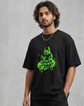 Person wearing a black t-shirt with green graphics and black cargo pants against a gray concrete wall.