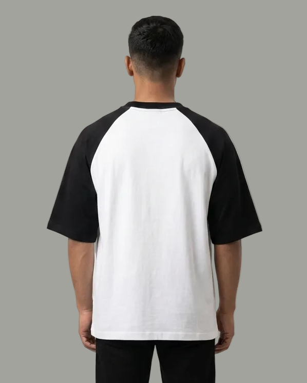 A man viewed from behind wearing a black and white raglan t-shirt, black pants, and white sneakers against a plain light gray studio background.