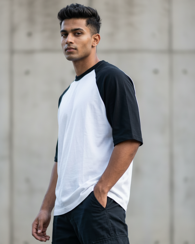 A man wearing a white and black raglan baseball t-shirt and dark cargo pants, standing against a textured concrete wall.
